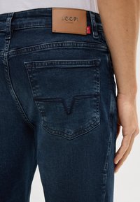 Jeans in denim blu scuro con una texture liscia. Presentano una toppa in pelle sulla vita con la dicitura "JOOP!" e una tasca posteriore con una cucitura a forma di V.