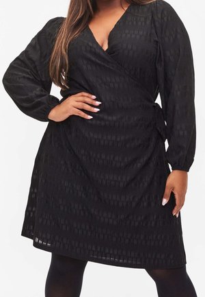 Femme portant une robe portefeuille noire texturée à manches longues et col en V, posant avec une main sur la hanche sur un fond blanc.