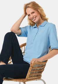 Hellblaues Poloshirt mit Kragen, drei Knöpfen und kurzen Ärmeln, kombiniert mit dunklen Jeans, sitzt in einem geflochtenen Stuhl.