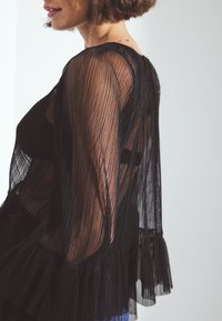Blusa nera plissettata in tessuto trasparente, con maniche lunghe e orlo a volant. La texture è leggera e scende fluida.