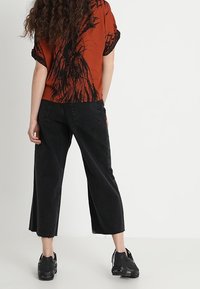 Röd t-shirt med kort ärm och svart grafiskt tryck, matchad med högmidjade svarta caprjeans. Svarta sneakers, enkel bakgrund.