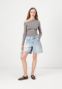 Haut gris à épaules dénudées à manches longues, shorts en denim bleu clair et ballerines en cuir léopard marron. Tient un sac à main argenté. Fond simple.
