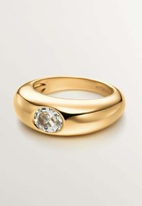 Anillo de oro con una superficie lisa y pulida y una gema ovalada clara incrustada en un lado. Diseño de banda circular, brillante y contemporáneo.