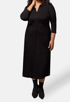 Kobieta ubrana w czarną midi sukienkę na guziki z rękawami do połowy łokcia i czarne botki, stojąca na prostym tle.