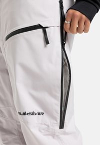 Lichtgrijze Quiksilver-broek met zwarte ritsen en gaasventilatie, close-up van de zijzak en een hand die de rits trekt.