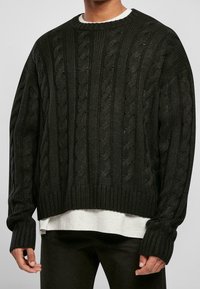 Suéter negro de punto trenzado con un corte holgado, puños y dobladillo acanalados. Presenta patrones texturizados y un cuello redondeado sobre una camisa blanca.