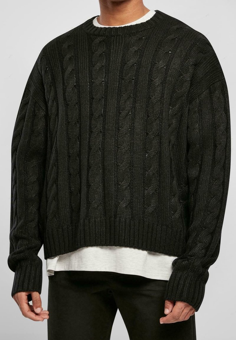 Suéter negro de punto trenzado con un corte holgado, puños y dobladillo acanalados. Presenta patrones texturizados y un cuello redondeado sobre una camisa blanca.