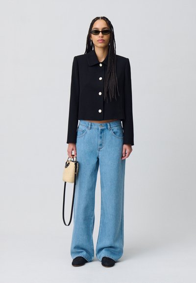 Veste noire courte avec de gros boutons blancs, jean large bleu clair, petit sac tissé et chaussures noires à enfiler. Des lunettes de soleil complètent le look.