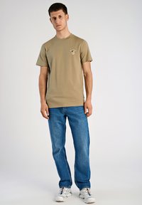Khaki bawełniany t-shirt z krótkim rękawem, okrągłym dekoltem i nadrukiem na piersi, zestawiony z niebieskimi dżinsami i białymi trampkami.