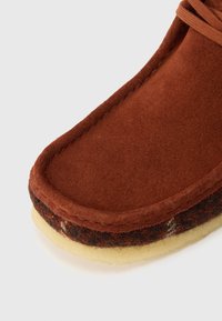Rudiamos suedes batai su apvaliu pirštu, storu kreminės spalvos tarpu, ir tamsiu tekstiliniu akcentu palei pagrindą. Matoma siuvinėta krašto dalis.
