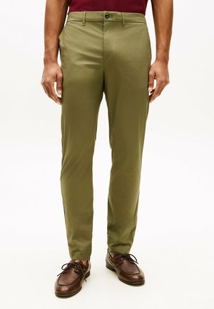 Mężczyzna w oliwkowo-zielonych obcisłych spodniach, brązowych skórzanych butach sznurowanych oraz w bordowej koszuli wpuszczonej do środka, stojący z rękami swobodnie opuszczonymi.