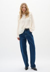 Blusa crema con dettagli a volant e pizzo, abbinata a jeans blu a vita alta e gamba dritta, e tacchi a punta nera.