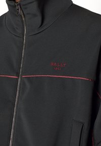 Giacca nera con colletto alto, chiusura con zip, cuciture rosse e logo ricamato "BALLY 1851" sul petto sinistro. Texture liscia e design aderente.