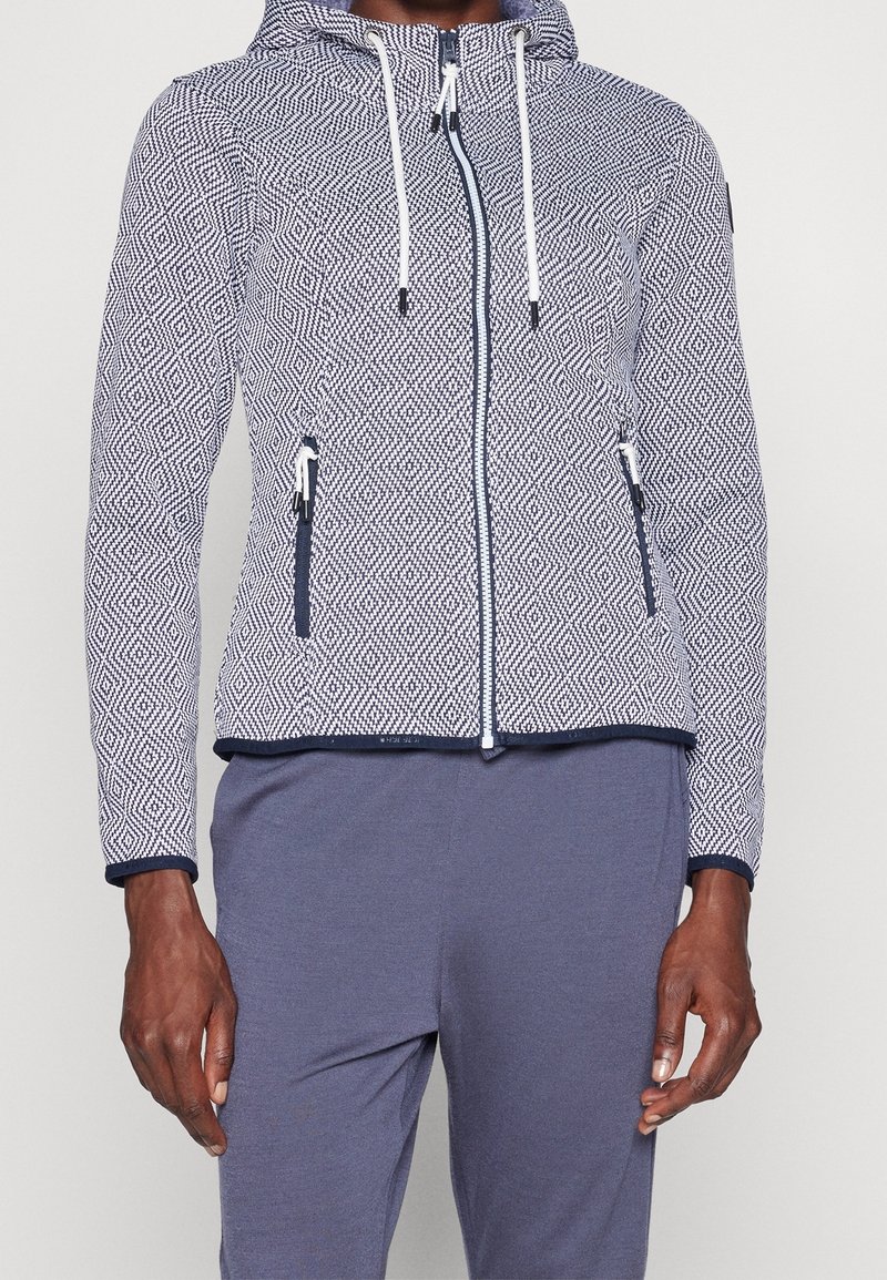 Homme portant un sweat à capuche zippé à motif avec cordons blancs et pantalon bleu marine, debout les bras détendus le long du corps.