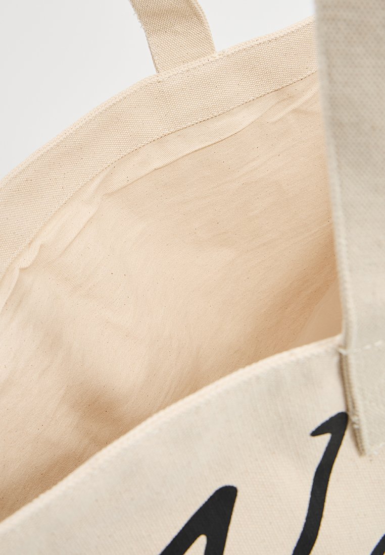 Gros plan sur un sac fourre-tout en coton naturel vide, montrant des poignées cousues et une partie d'un motif imprimé noir sur le tissu extérieur.