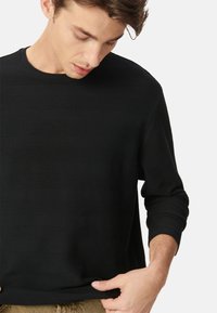 Uomo che guarda in basso, indossando una camicia nera a maniche lunghe con texture e pantaloni marrone chiaro, mentre tiene l'orlo della camicia.