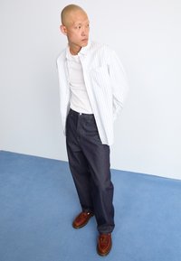 White striped button-up shirt over a plain white tee, paired with dark blue loose jeans and brown lace-up shoes. Light background.