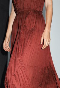 A hand gently holds the flowing fabric of a deep red, pleated dress against a neutral background, showcasing texture and elegance.
