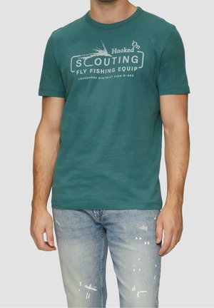 Homme portant un t-shirt bleu sarcelle avec le texte "Scouting Fly Fishing Equip" et un jean bleu clair usé sur fond uni.