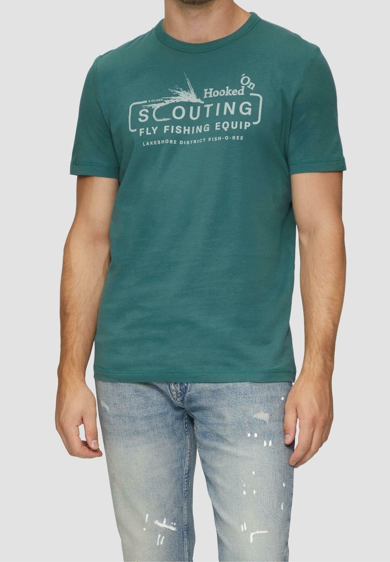 Homme portant un t-shirt bleu sarcelle avec le texte "Scouting Fly Fishing Equip" et un jean bleu clair usé sur fond uni.