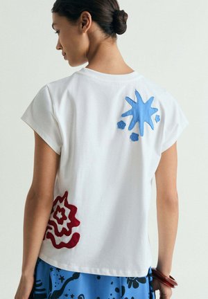 Mujer con el pelo recogido en un moño que lleva una camisa blanca de manga corta con parches abstractos azules y rojos en la espalda y una falda azul con estampado.