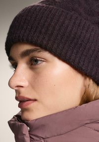 Sötét lila bordázott kötött sapka egy személyen, világos rózsaszín pufi kabáttal párosítva. Sima bőr finom szeplőkkel és természetes sminkkel.