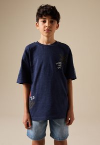 Chłopiec stojący w granatowym T-shircie z napisem "Under the sky" i grafiką palmy, w połączeniu z jasnymi jeansowymi szortami.