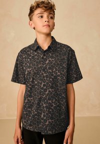 Un adolescent portant une chemise à manches courtes, boutonnée, avec un imprimé léopard noir et brun foncé, se tenant devant un fond beige uni.
