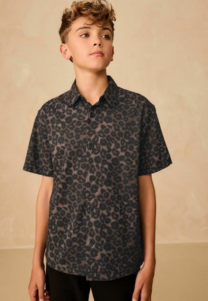 Un adolescent portant une chemise à manches courtes, boutonnée, avec un imprimé léopard noir et brun foncé, se tenant devant un fond beige uni.