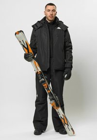 Schwarze Skijacke mit Kapuze, schwarze Skihose, schwarze Handschuhe und schwarze Stiefel. Hält grau-orangefarbene Skier mit schwarzen Akzenten.