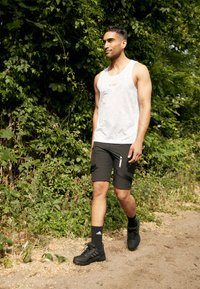 Weißes Mesh-Trägershirt mit orangefarbenem Logo, schwarze Shorts mit Reißverschlüssen, schwarze Sportschuhe, die auf einem Weg gehen, umgeben von Grün.