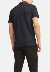 Polo shirt gris foncé à manches courtes, avec col et fentes sur les côtés, associé à un pantalon noir. Le tissu semble lisse et léger.