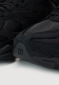 Chaussures de course noires présentant un mélange de matériaux en daim et en maille, avec une semelle en caoutchouc texturé et un notable logo "N" à l'arrière.