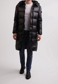 Černá péřová bunda s kapucí, prošívaný vzor, přední zip a knoflíky. Nositelná přes bílou košili, doplněná tmavými džínami a černými botami.