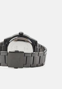 Schwarze Edelstahl-Uhr mit einer glatten, gebürsteten Oberfläche, rundem Zifferblatt, metallischem Verschluss und sichtbarem Uhrwerk auf der Gehäuserückseite.