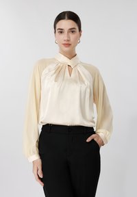 Kvinde med mørkt hår iført en cremefarvet silkebluse med nøglehulssnit og sorte bukser, stående mod en ensartet lys baggrund.