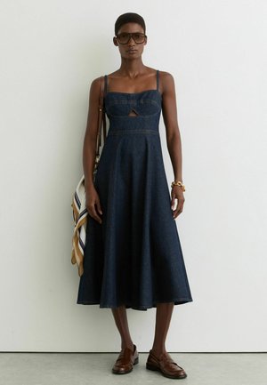 Vrouw draagt een midi-jurk van donker denim met dunne bandjes, bruine loafers, oversized zonnebril, gouden armband en draagt een tas met een gestreepte sjaal.