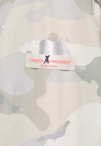 Tkanina w wzór moro w jasnoszarym i beżowym kolorze o gładkiej fakturze. Posiada białą metkę z napisem "FRIEDA FREDDIES NEW YORK" w kolorze różowym oraz małą ikoną psa.