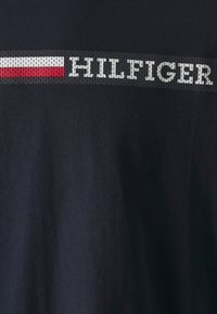 Tricou din material negru, cu un logo „HILFIGER” proeminent de culoare gri, însoțit de o bandă orizontală roșie și albă deasupra. Textură netedă, design casual.