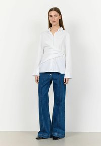 Camicia bianca a maniche lunghe con un design attorcigliato in vita, abbinata a jeans blu a gamba larga con cuciture a contrasto e un dettaglio con bottone.
