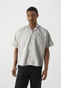 Kurzarmhemd in hellgrauem Stoff mit subtiler Textur. Verfügt über eine Brusttasche und einen abgerundeten Kragen.