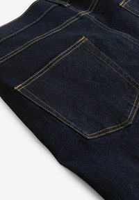 Tumma farkut, joissa on yksi takatasku, jossa on terävä muotoilu ja kontrastivärinen ommel. Materiaali on sileä.