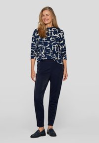 Marineblaues Langarmoberteil mit weißem abstraktem Muster, kombiniert mit dunklen, maßgeschneiderten Hosen. Das Model trägt schwarze Slipper und steht vor einem neutralen Hintergrund.