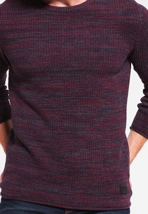 Mężczyzna w teksturowanym, ciemnoczerwonym i granatowym swetrze z długimi rękawami, z rękami spoczywającymi za plecami.