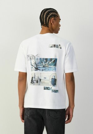 Personne aux cheveux tressés portant un t-shirt blanc avec des illustrations de science-fiction bleutées dans le dos et un jean noir.
