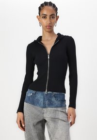 Svart ribbestrikket zip-up hettegenser med en tettsittende silhuett. Kombinert med høy midje, to-toners denimjeans med en teksturert overflate.