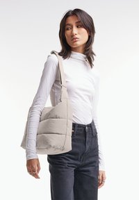 Borsa a spalla beige con spalline voluminose, con un design trapuntato e una tracolla larga, indossata da un modello che indossa un dolcevita bianco e jeans scuri.