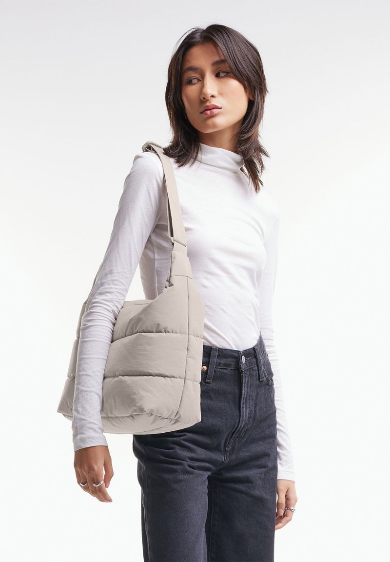 Borsa a spalla beige con spalline voluminose, con un design trapuntato e una tracolla larga, indossata da un modello che indossa un dolcevita bianco e jeans scuri.