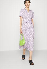 Vestido floral lilás, com mangas curtas, fecho à frente em botões, cintura cingida, e bainha com rufos. Uma bolsa verde e mules pretas de salto complementam o conjunto.