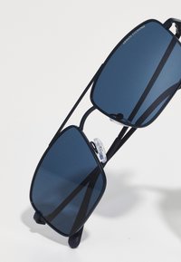 Ochelari de soare dreptunghiulari de culoare albastru marin, cu lentile întunecate, ramă din metal și suporturi nazale transparente. Au un design minimalist și o structură cu pod dublu.